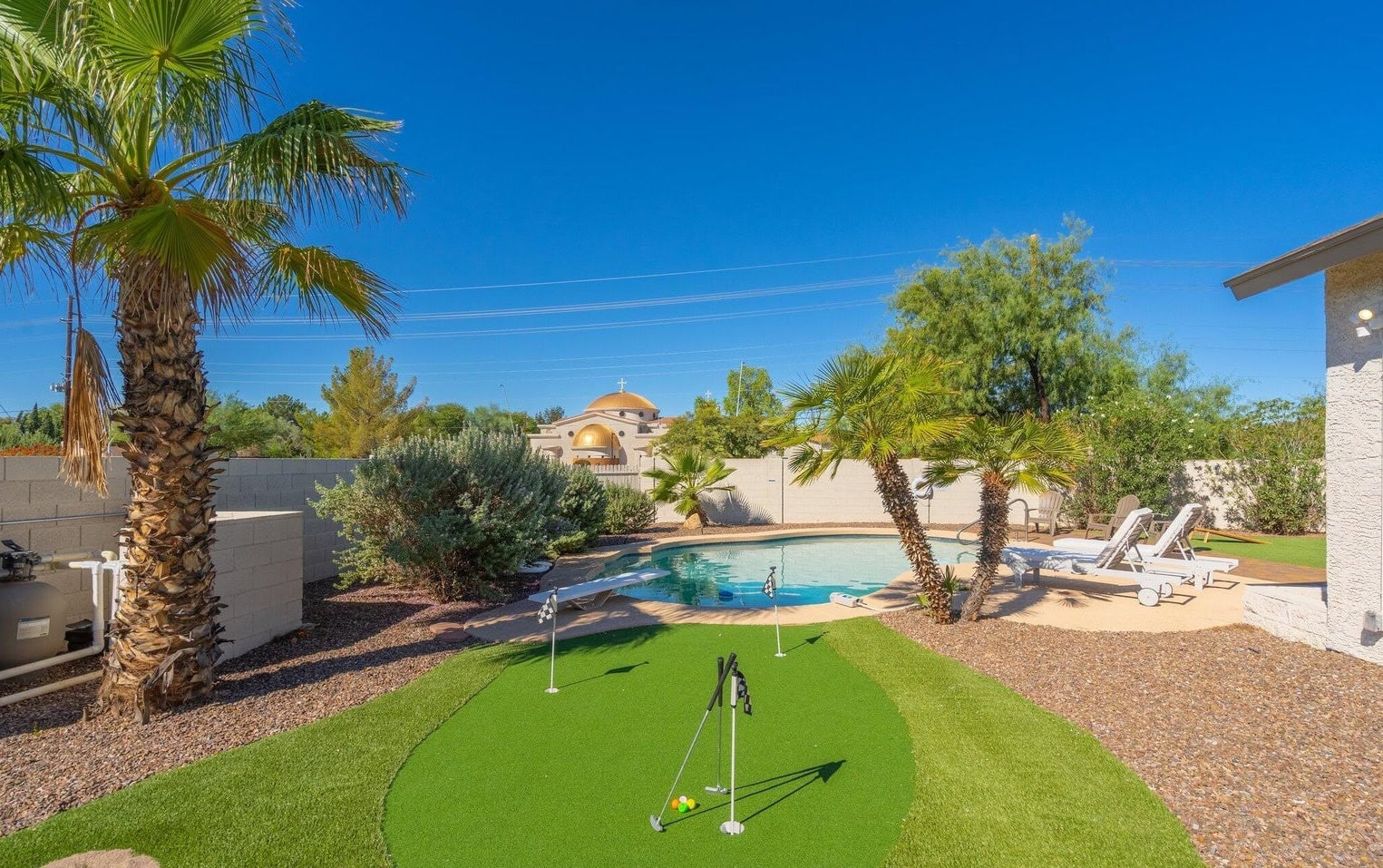 A backyard with palm trees, a small artificial putting green, and a swimming pool surrounded by lounge chairs, under a clear blue sky.