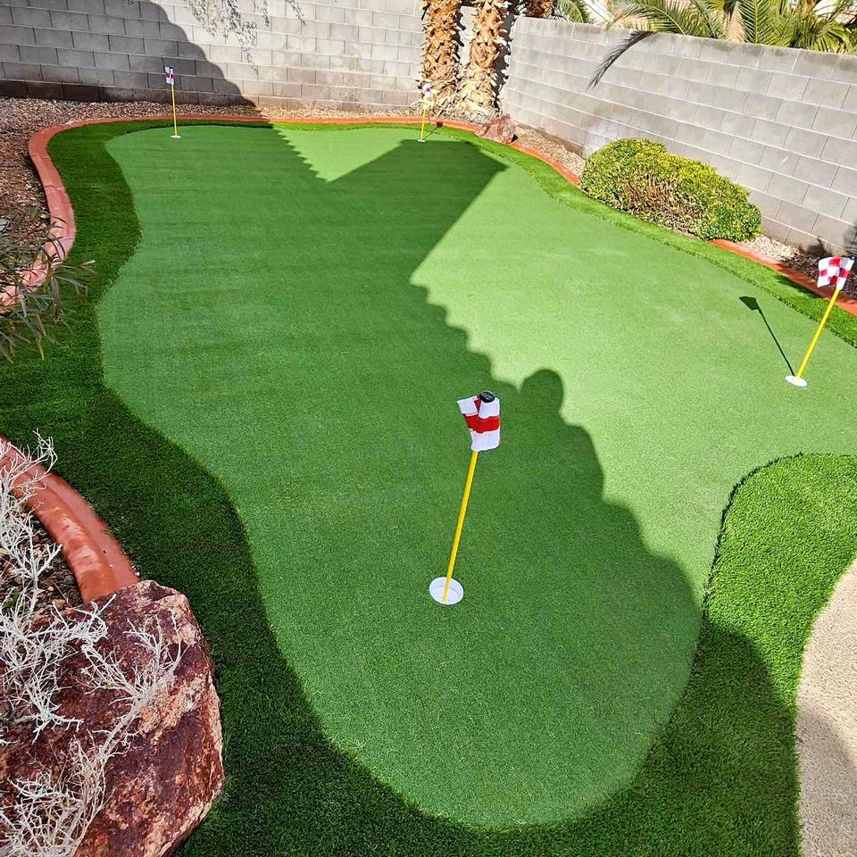 A backyard putting green with three putting holes, each marked by a red and white flag, is surrounded by a brick border and landscaping with rocks and desert plants.
