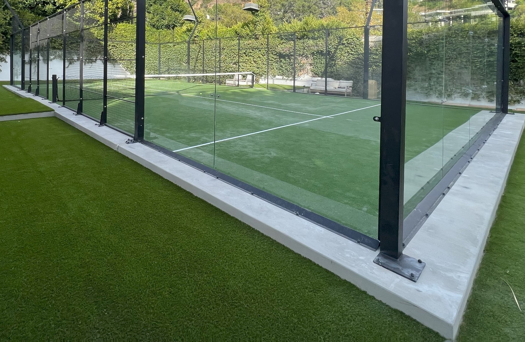 A fenced padel court with glass walls and an artificial grass surface by AZ Turf and Pavers, surrounded by green hedges and trees. Benches are visible outside the court.