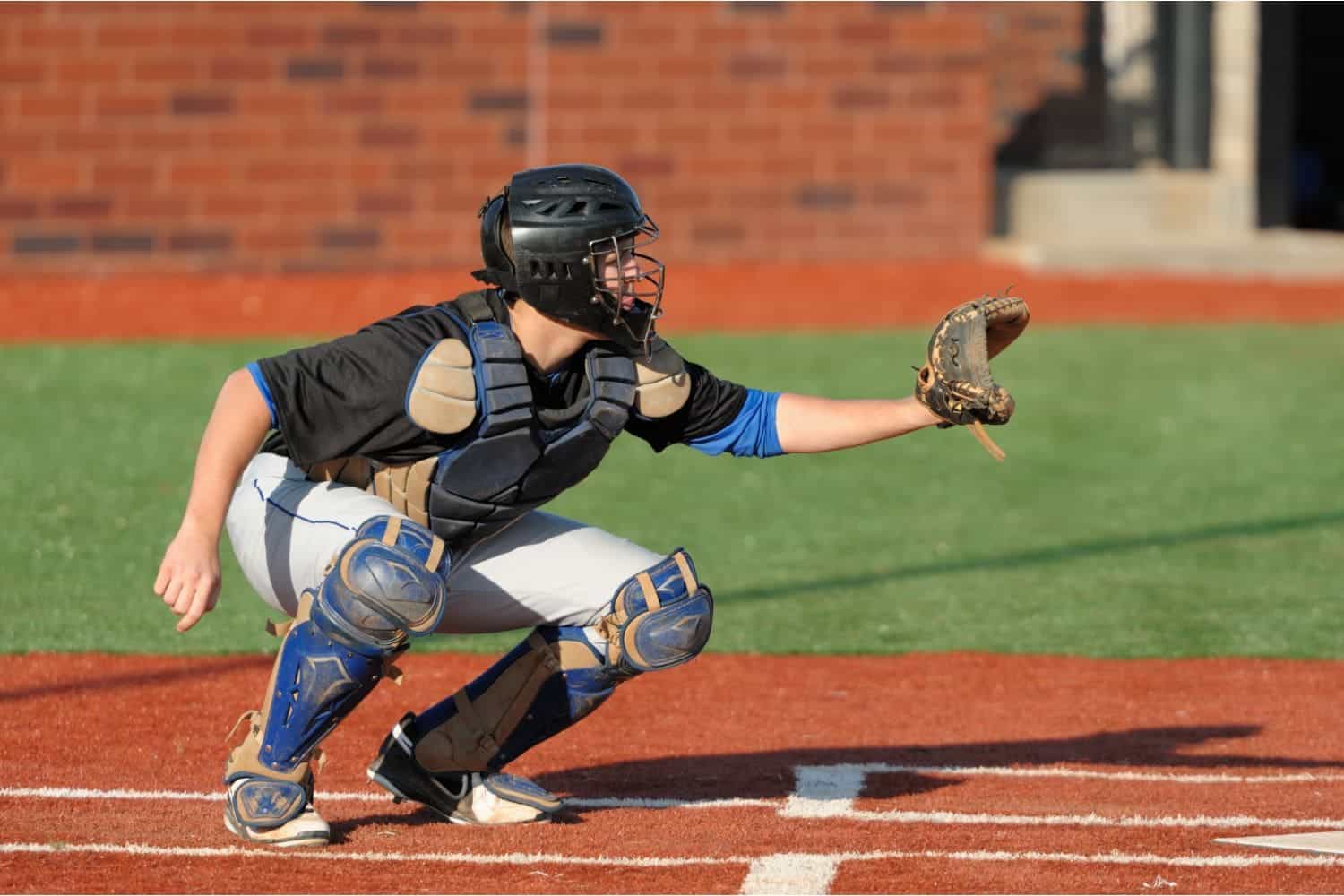 A baseball catcher in full gear crouches behind home plate on a pristine AZ Turf and Pavers baseball field, wearing a helmet, chest protector, and holding a glove out to catch a pitch.