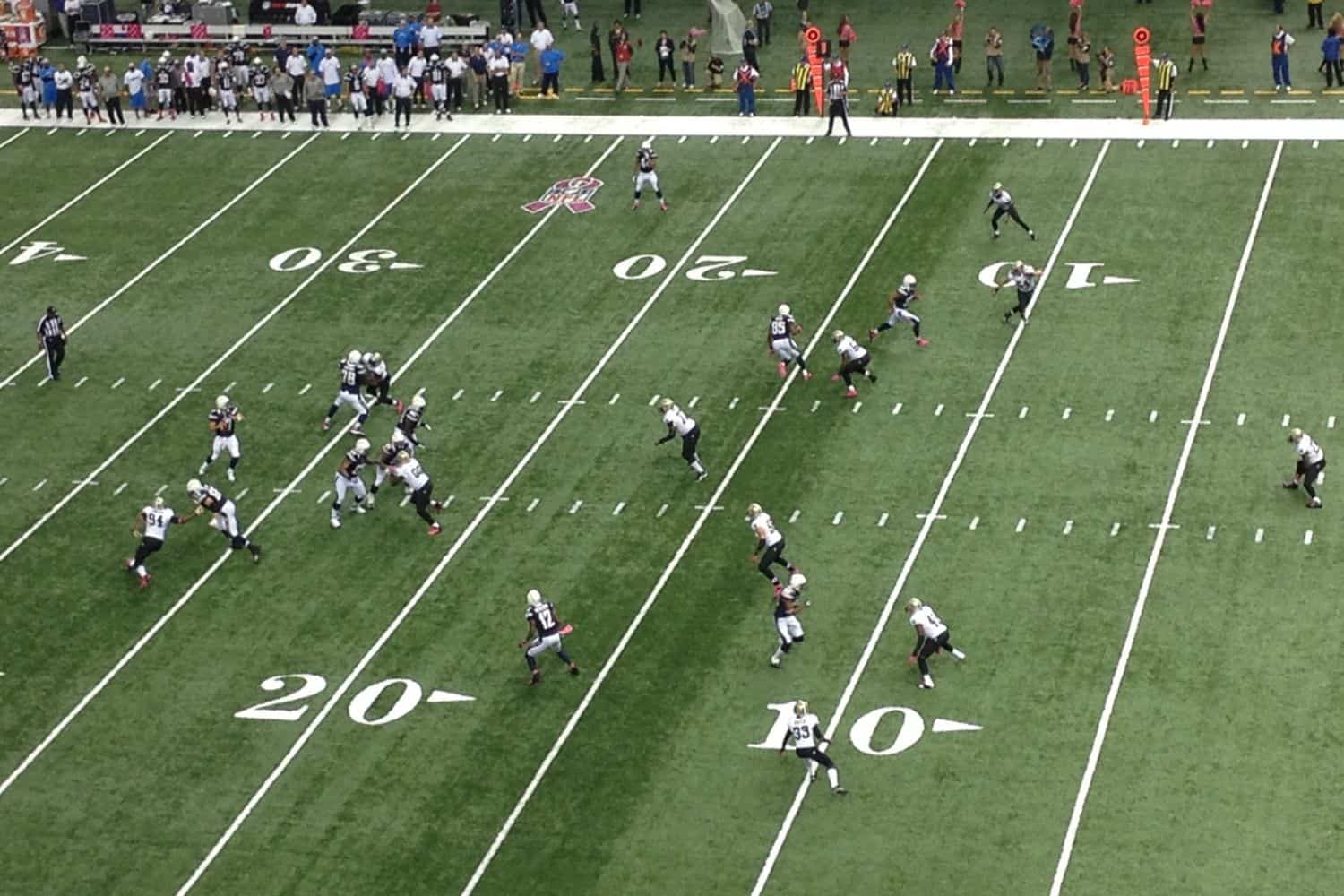 A high-angle view of an American football game showcases players lined up on the field near the 20-yard line, with offensive and defensive formations visible on the well-maintained AZ Turf and Pavers surface as spectators and staff watch from the sidelines.