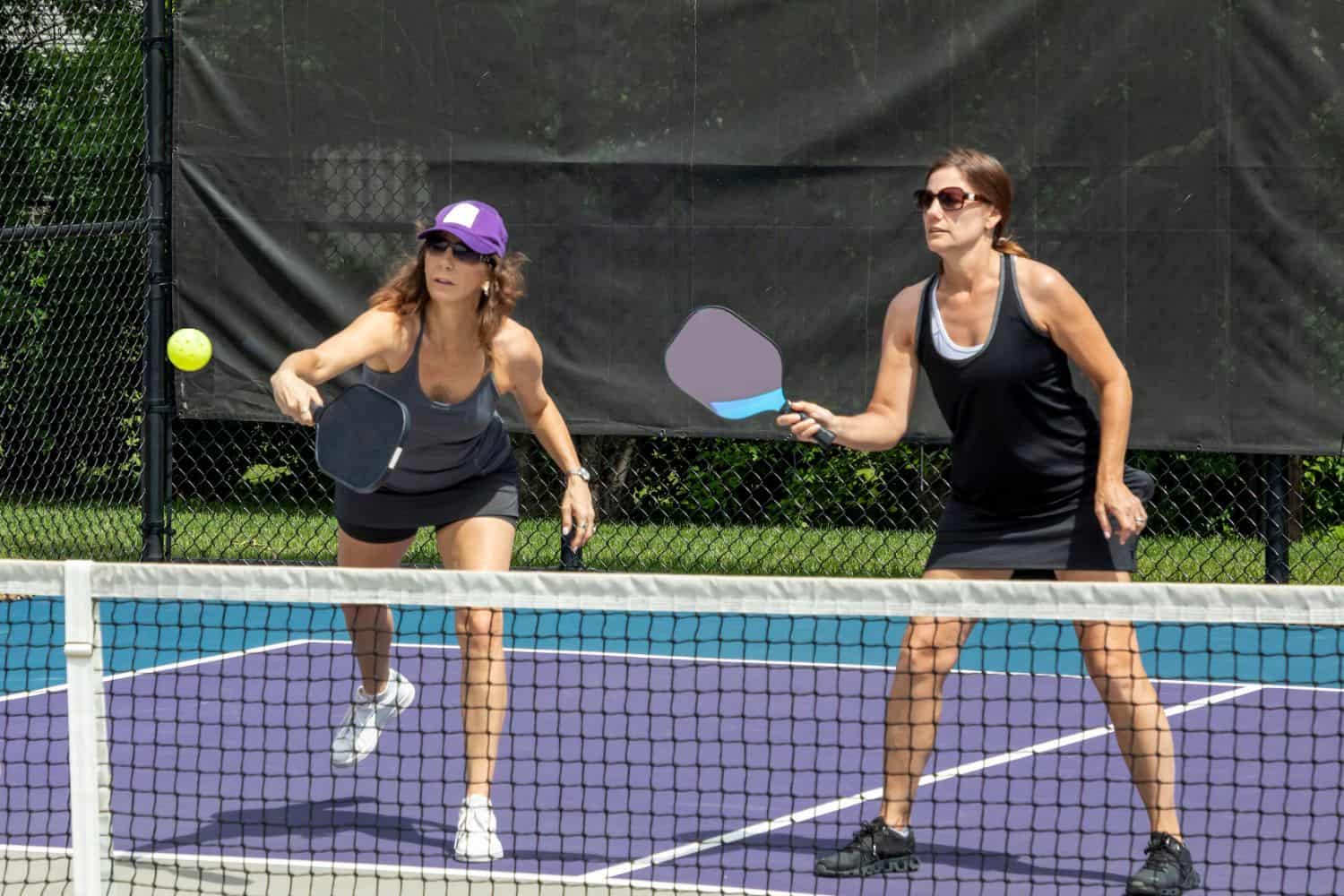 Two women playing pickleball on an outdoor court, both holding paddles and preparing to hit the ball atop pristine AZ Turf and Pavers, with one stepping forward and the other standing ready behind her.