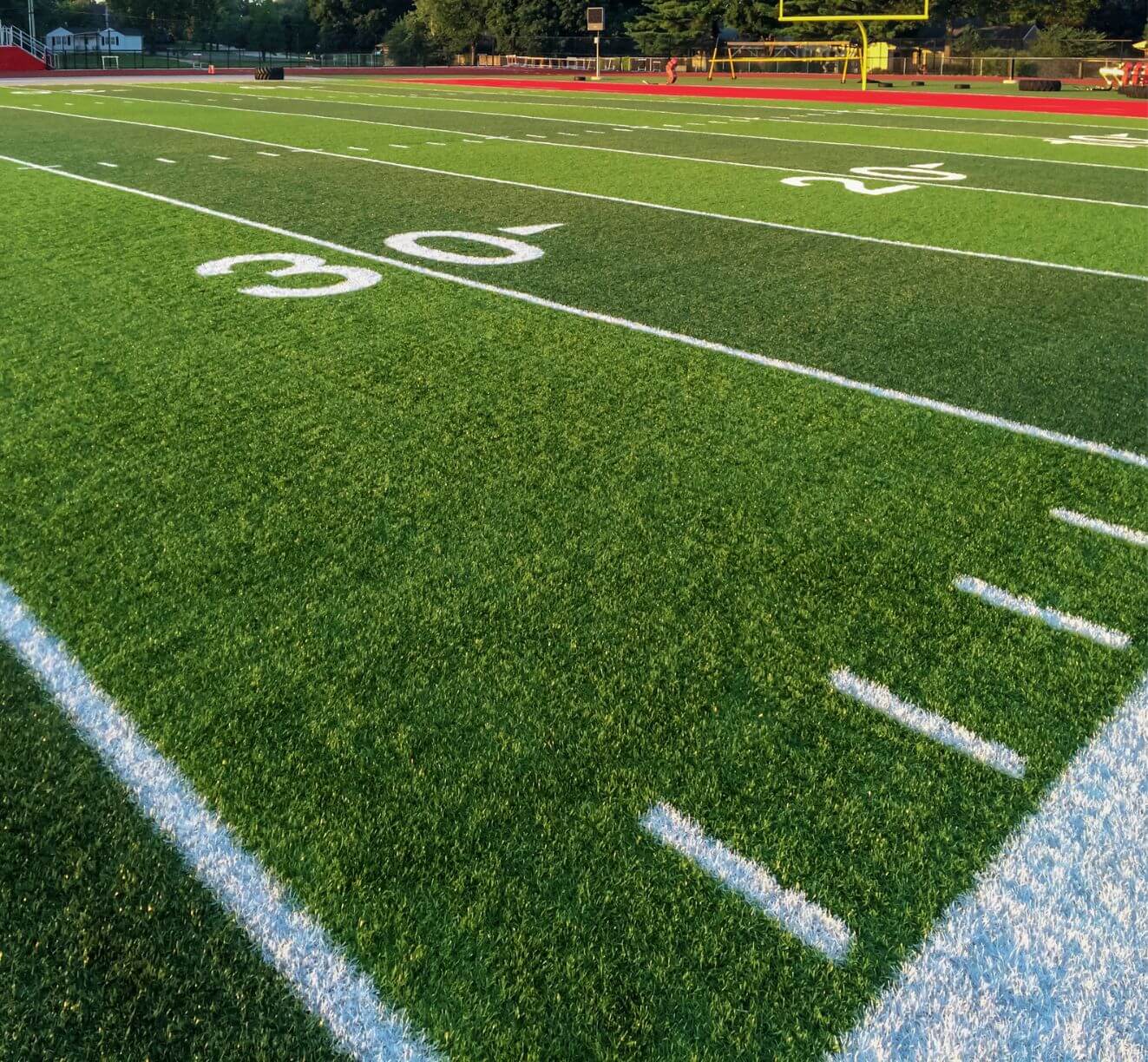 A close-up view of a football field by AZ Turf and Pavers shows the 30-yard line, crisp white yard markings, green artificial turf, and a yellow goalpost in the background under bright sunlight.