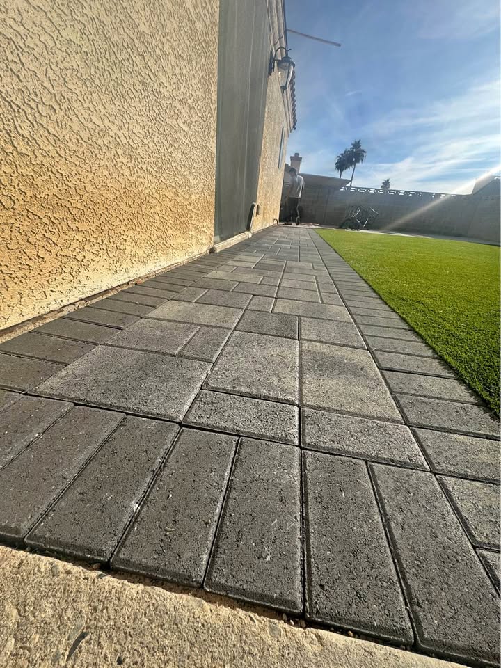 A paved walkway with rectangular gray bricks runs alongside a textured beige exterior wall, next to a strip of artificial green grass, under a clear, sunny sky.