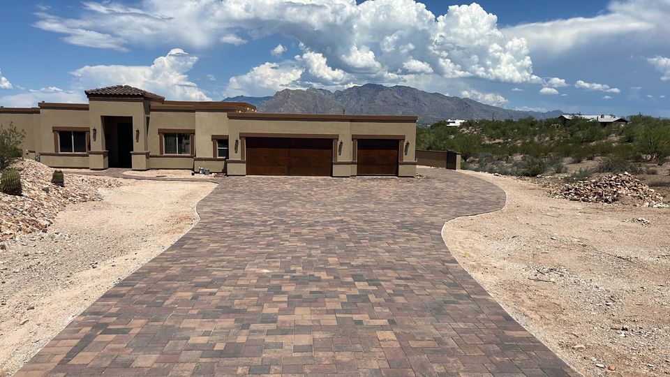 Single-story modern house with a three-car garage, tan exterior, and a long, wide brick driveway. The house is surrounded by desert landscape and mountains in the background under a partly cloudy sky.