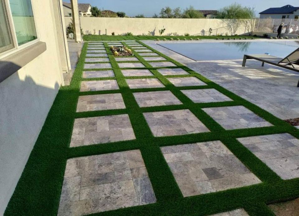 A modern backyard features a swimming pool and a walkway of large square stone tiles set in a grid pattern with green artificial grass in between. The scene is bright and well-maintained, with lounge chairs by the pool.