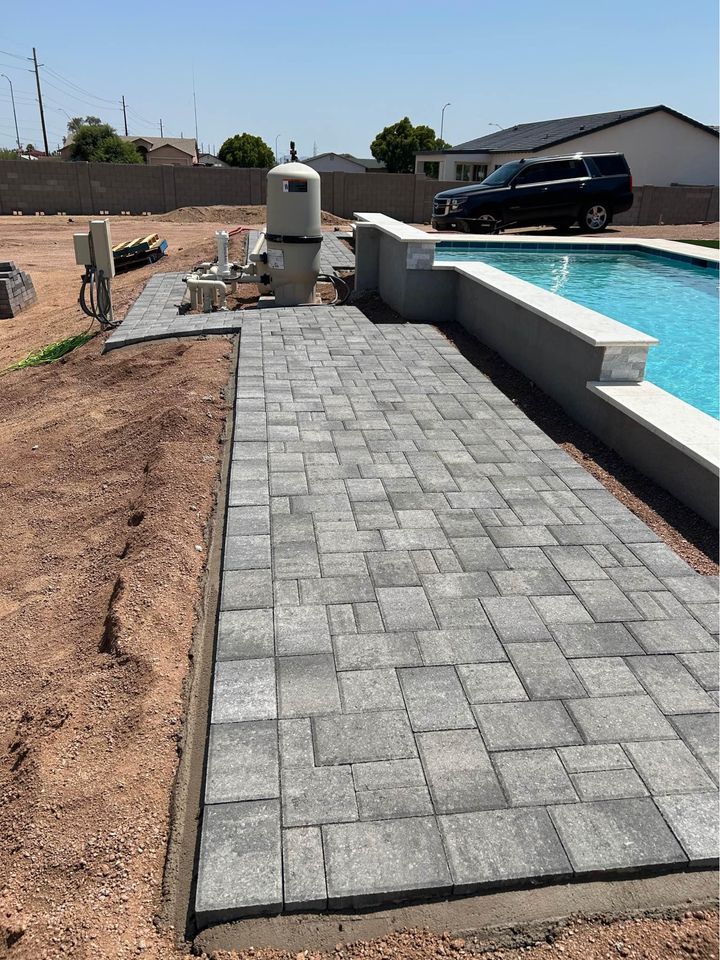 A freshly paved gray stone walkway runs alongside a backyard swimming pool, bordered by unfinished landscaping with dirt and construction materials nearby. A black SUV is parked in the background.