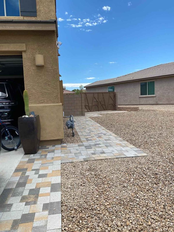 A paved walkway bordered by rocks leads to a wooden gate between two beige houses. A potted cactus and a decorative bird sculpture are near a garage on the left. The sky is blue with a few clouds.