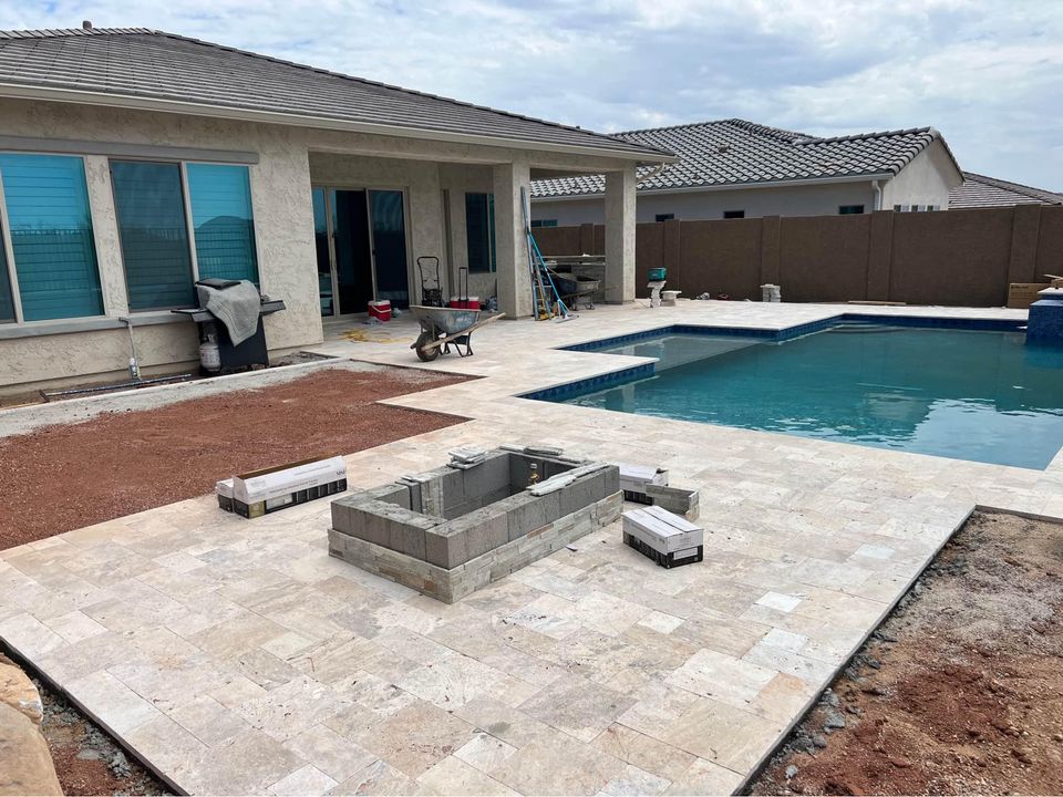 A backyard under construction with a tiled patio, an unfinished rectangular fire pit, stacked bricks, a wheelbarrow, and a pool next to a house with large windows.