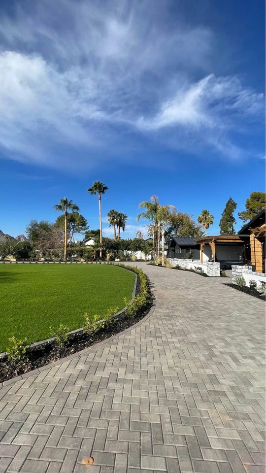A paved pathway curves alongside a neatly trimmed green lawn with palm trees and shrubbery, leading to modern houses under a bright blue sky with wispy clouds.