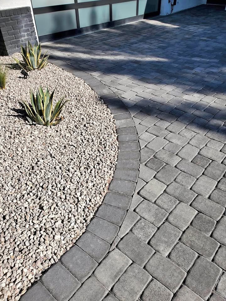 Curved border of gray stone pavers meets a bed of light-colored gravel with a few spiky green plants, in front of a modern garage door and driveway.