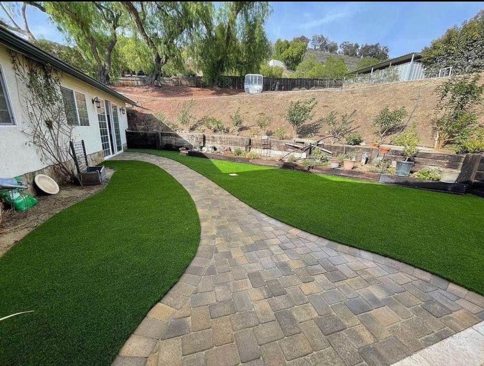 A paved walkway curves through a backyard with artificial grass on both sides, next to a house. The yard is bordered by a sloped hill with plants, trees, and a few garden beds.
