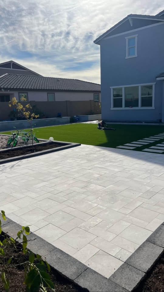 A backyard with a newly paved patio, artificial grass lawn, stepping stones, and a modern gray house on the right under a partly cloudy sky. A small green bike and garden hose are visible on the grass.