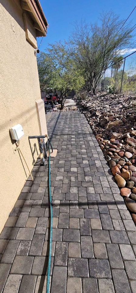 A paved walkway made of rectangular gray stones runs alongside a tan house, with a green garden hose lying across it. Rocks and desert plants line the right side under a clear blue sky.