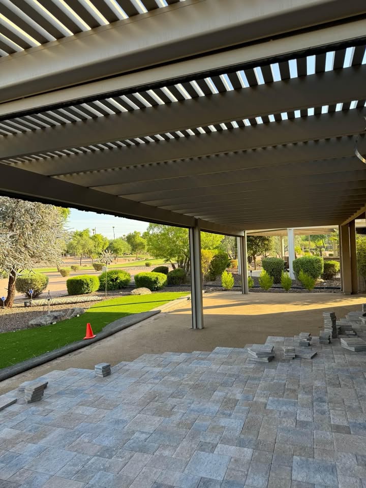 View of a partially covered patio with tile flooring being installed, stacks of tiles, and a landscaped garden with bushes and trees in the background under a clear sky.